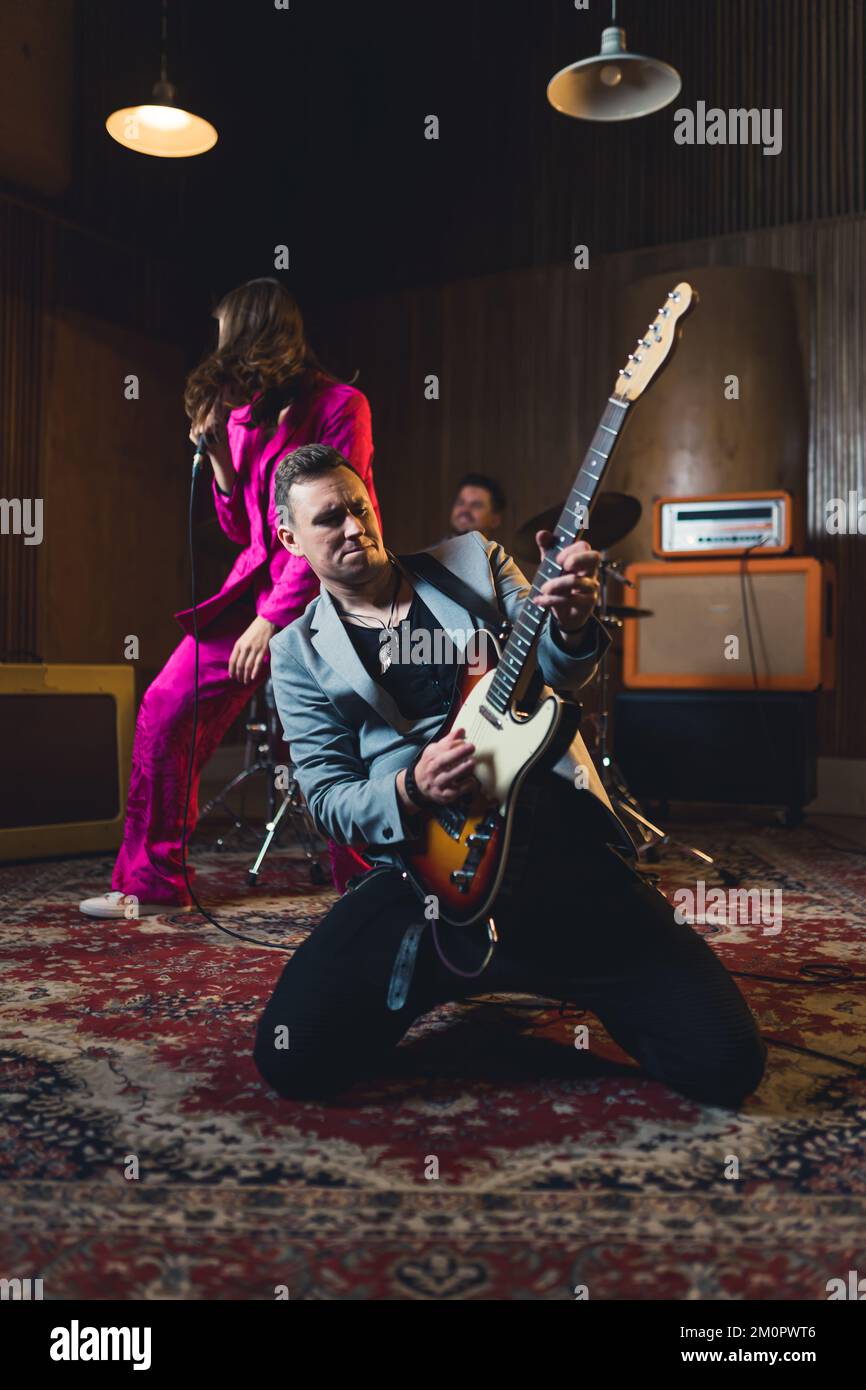 Ein Mann, der auf Knien Gitarre spielt, mit einer Musikband in einem Studio auftritt. Hochwertiges Foto Stockfoto