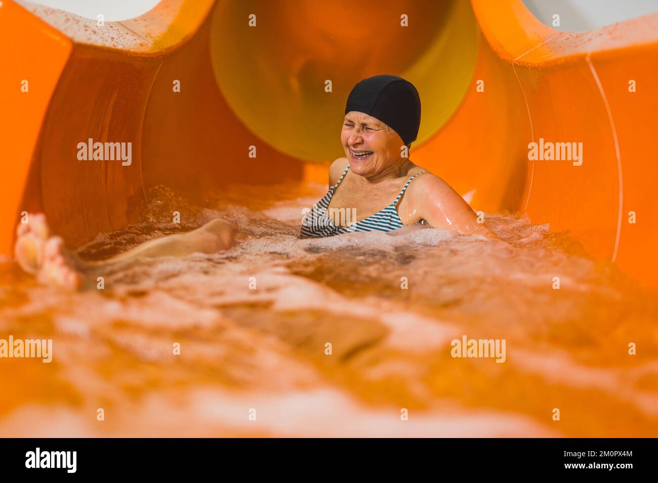 Amüsierte ältere, lachende Erwachsene Frau in gestreiftem Badeanzug und schwarzer Haarkappe, die auf einer orangefarbenen Wasserrutsche gleitet. Aktive und unterhaltsame Aktivitäten. Hochwertiges Foto Stockfoto