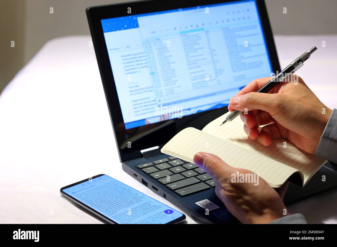 Eine Nahaufnahme der Hände einer Person, die einen Bleistift und ein Notizbuch hält und Informationen aus Dokumenten aufschreibt, die auf einem Laptop-Computer angezeigt werden. Stockfoto