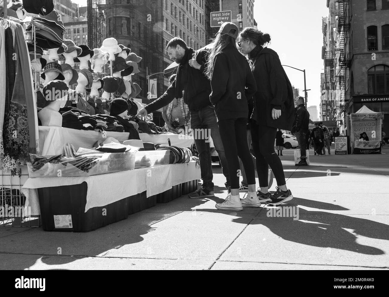 Eine Gruppe junger Leute, die Kappen und Hüte von Straßenverkäufern auf dem Bürgersteig in Manhattan, New York City, kaufen. Schwarz-weiße Straßenfotografie. Stockfoto