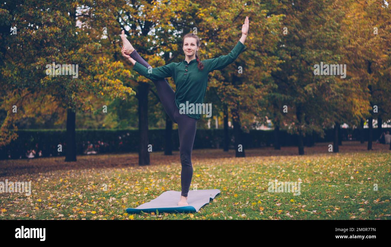 Ein lächelndes Mädchen professionelle Yoga-Lehrerin macht Balanceübungen, während sie auf einem Bein auf einer Matte auf grünem und gelbem Gras im Park steht. Die wunderschöne Herbstlandschaft ist sichtbar. Stockfoto