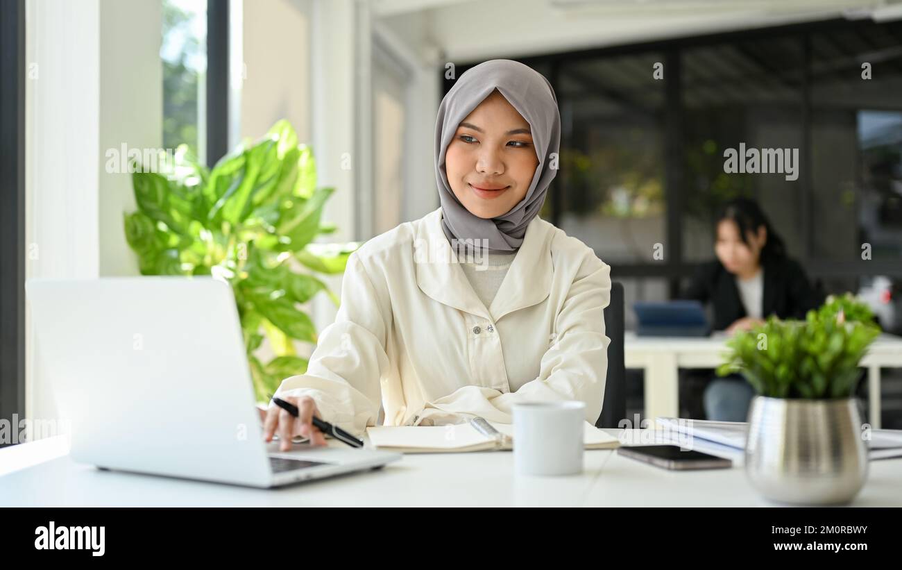 Wunderschöne, junge asiatische, muslimische Geschäftsfrau, die Hijab trägt, einen Laptop benutzt, im Büro arbeitet. Stockfoto