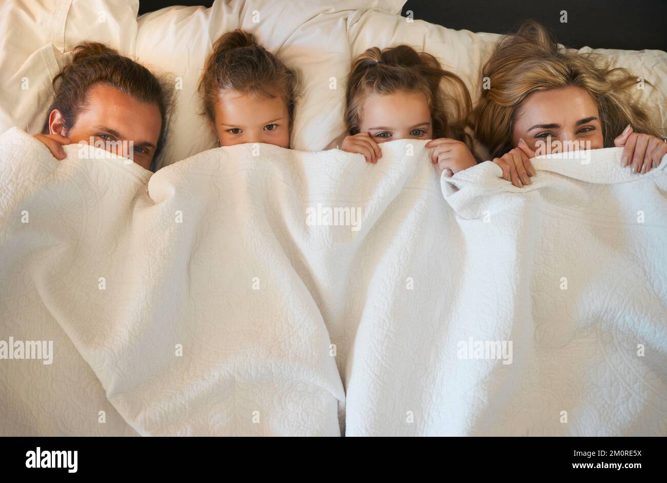 Porträt einer jungen, gemütlichen, weißen Familie, die in einem Bett zusammen unter einer Decke ruht. Gemütliche kleine Mädchen, die sich mit ihren Eltern im Bett entspannen Stockfoto