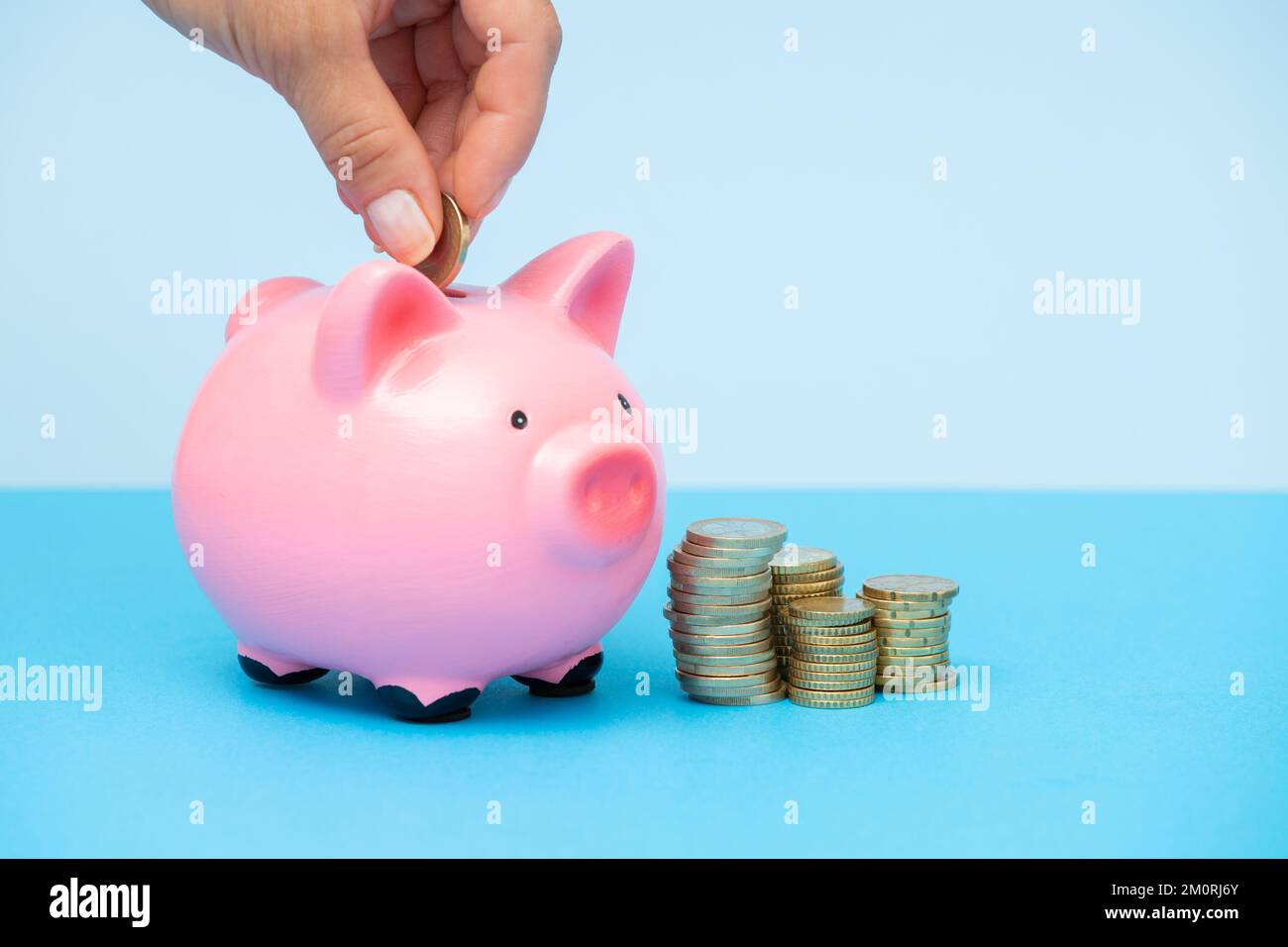 Person legt Geld in ein rosa Sparschwein auf einem blauen Hintergrund.Frauen Hände legen Münzen in eine Geldbox.Sparschwein mit Stapel von Münzen.das Konzept der Stockfoto