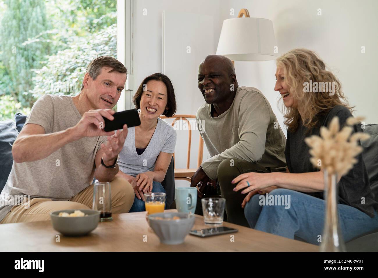 Senior man sharing pictures on smart phone with friends while sitting on sofa Stockfoto