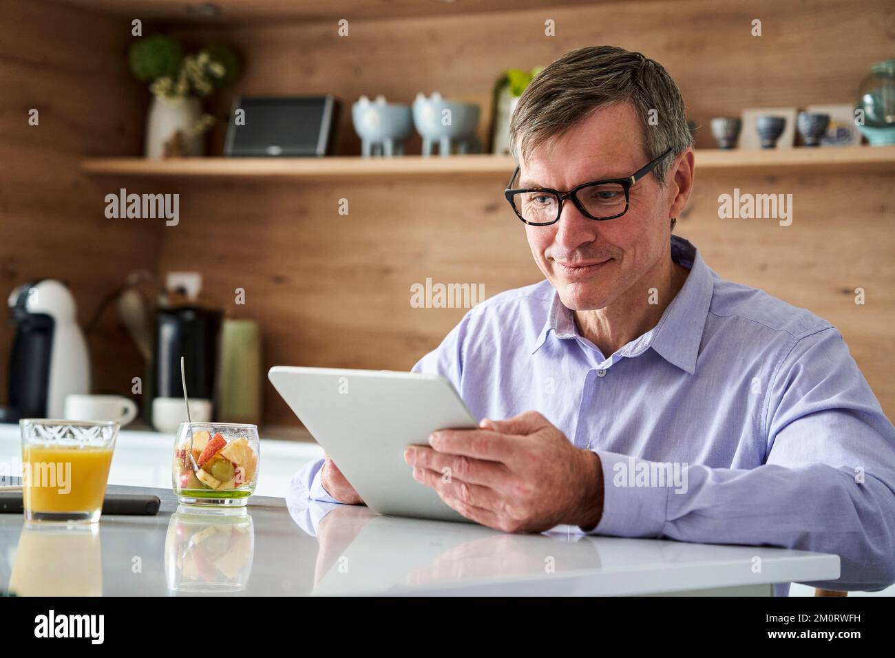 Professioneller Mann mittleren Alters, der lächelt und Nachrichten auf einem digitalen Tablet liest, während er zu Hause auf der Küchentheke frühstückt Stockfoto