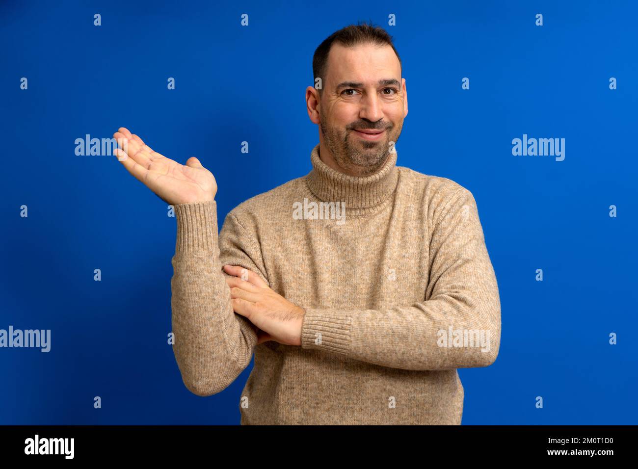 Lateinamerikanischer Mann mit Bart und braunem Rollkragenpullover in skeptischer Einstellung, der etwas in seiner Handfläche anbietet, isoliert auf blauem Hintergrund Stockfoto