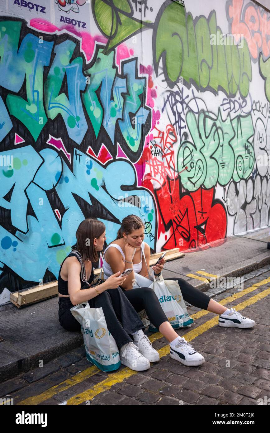 Zwei junge Erwachsene Frauen mit iphones, die auf dem Bordstein unter Graffiti sitzen Stockfoto