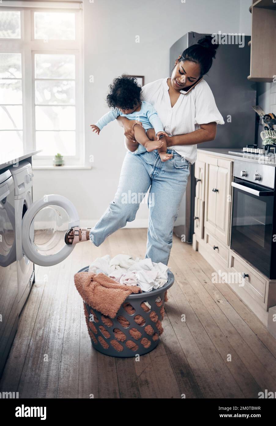 Babys Wäsche kann nicht warten. Eine junge Mutter, die ein Handy benutzt, während sie Hausarbeit macht und ihr Baby zu Hause hält. Stockfoto