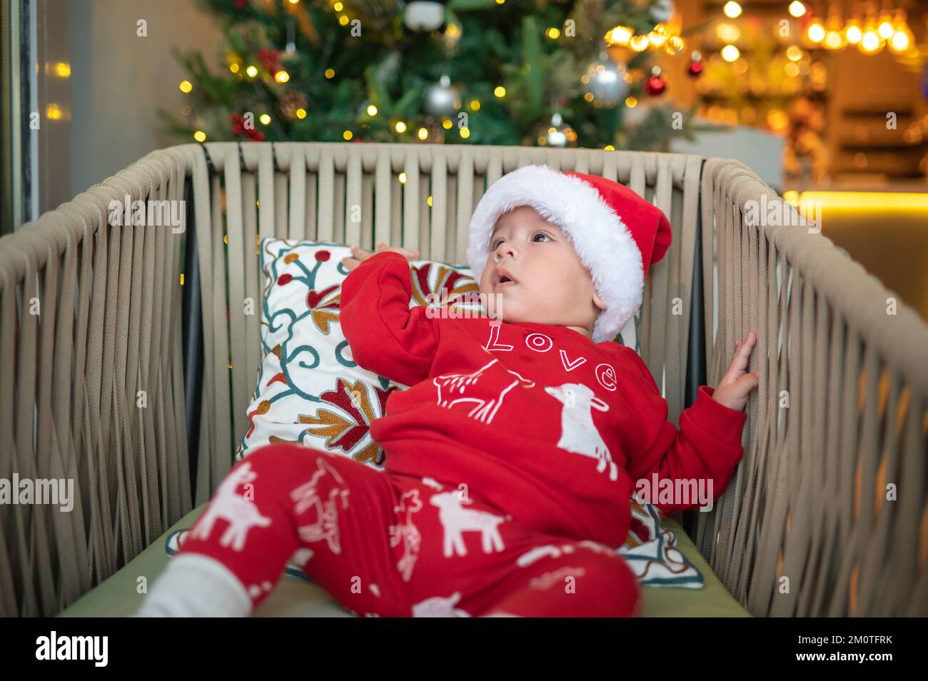 Süßes neugeborenes Kind mit Weihnachtsmannmütze und festlichem Outfit neben einem Weihnachtsbaum mit festlicher Dekoration und Fährenbeleuchtung für Neujahr und Weihnachten Stockfoto