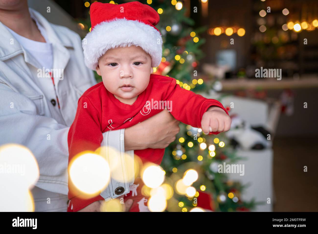 Süßes neugeborenes Kind mit Weihnachtsmannmütze und festlichem Outfit neben einem Weihnachtsbaum mit festlicher Dekoration und Fährenbeleuchtung für Neujahr und Weihnachten Stockfoto
