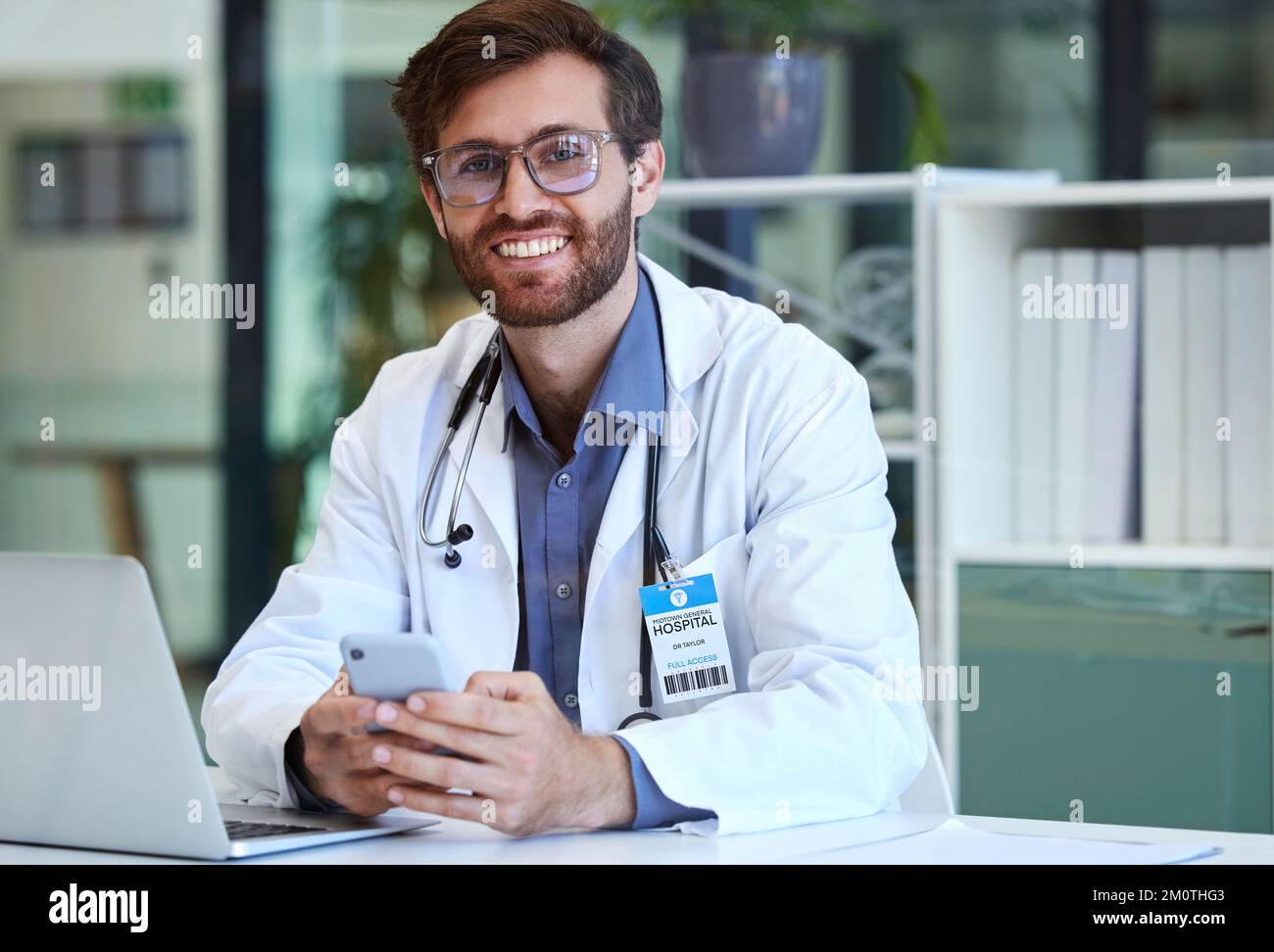 Arzt, Schreibtisch und Telefon mit Lächeln für SMS, Kommunikation oder Chats und gute Verbindung im Krankenhaus. Glücklicher Mann im Gesundheitswesen oder Mediziner Stockfoto