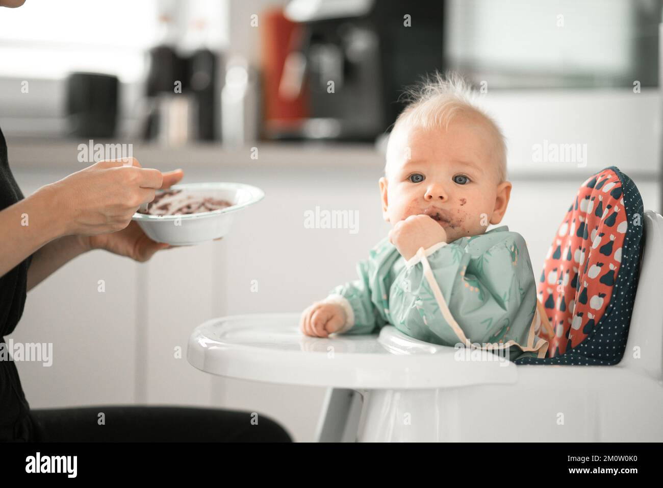 Mutter Löffel Fütterung ihres kleinen Jungen Kind in Babystuhl mit Fruchtpüree in der Küche zu Hause. Baby feste Nahrung Einführung Konzept Stockfoto