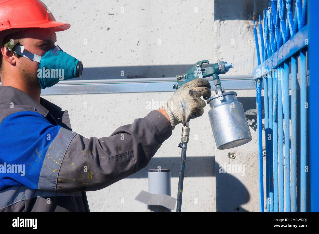 Industrieller Maler in Atemschutzgeräten und Helm lackiert Metallzaun aus der Sprühpistole. Zaun streichen am Sommertag. Authentischer Workflow. Berufsarbeiter im Overall. Stockfoto