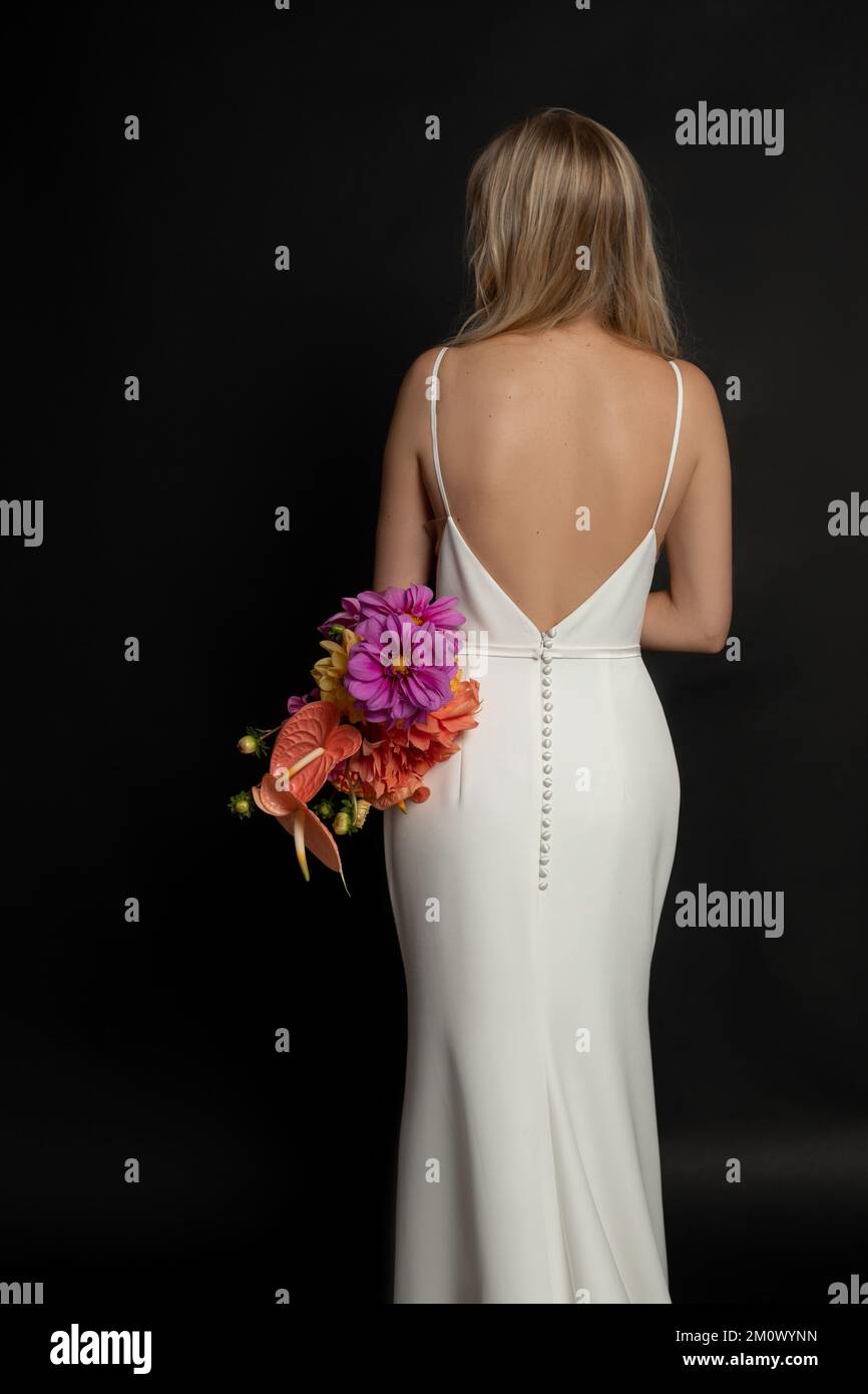 Senkrechte Rückansicht einer blonden Frau mit langen Haaren in weißem geknöpftem Kleid hält einen Haufen bunter Blumen in schwarzem Studio Stockfoto