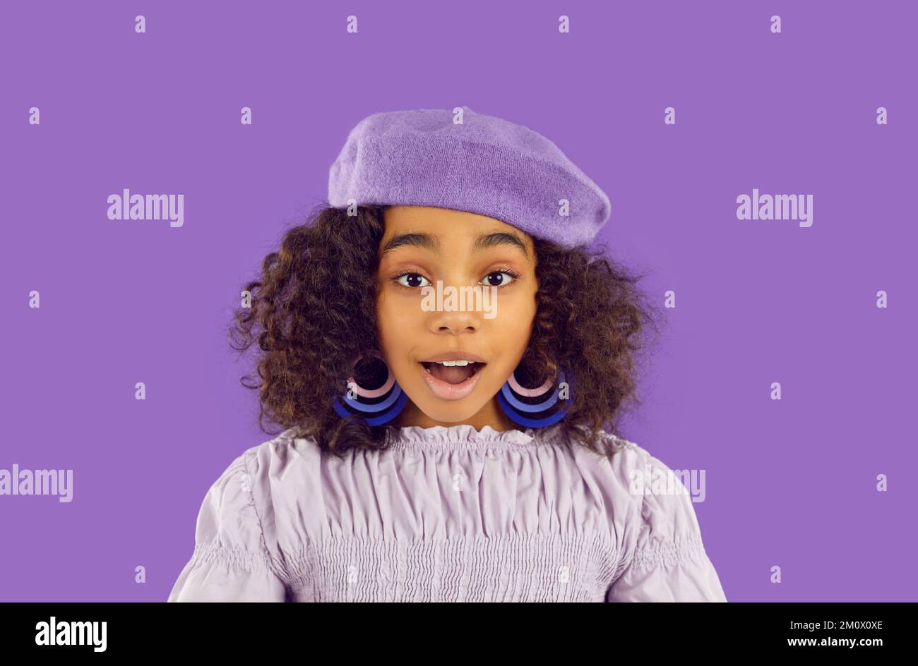 Schwarzes Mädchen in lila Baskenmütze und Ohrringen, die mit überraschtem Gesichtsausdruck auf die Kamera schaut Stockfoto