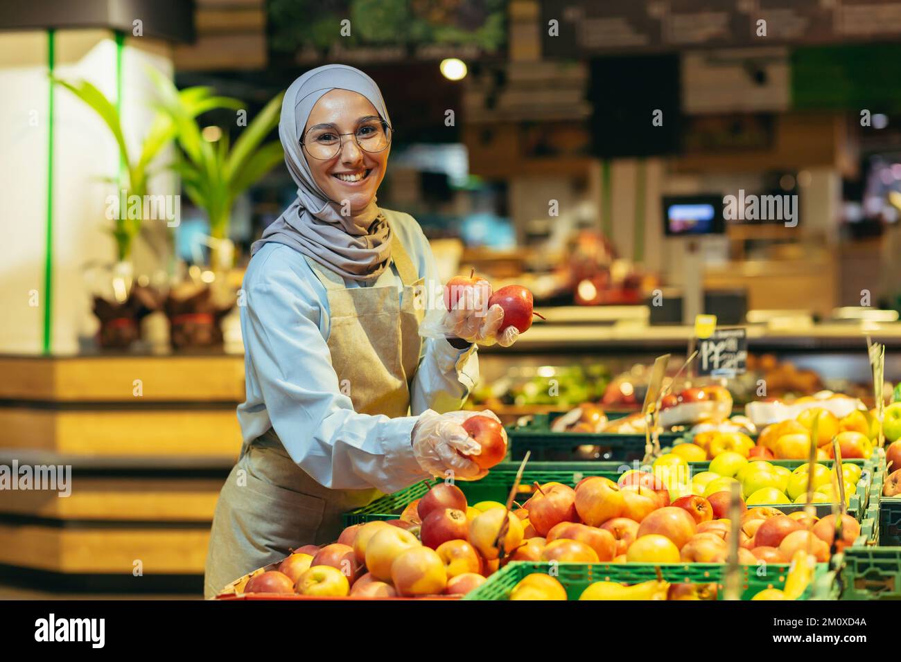 Eine junge, wunderschöne Hidschabi-Muslime, eine Supermarktkraft, legt Früchte in der Abteilung. Er hält Äpfel in den Händen, sieht in die Kamera, lächelt. Stockfoto