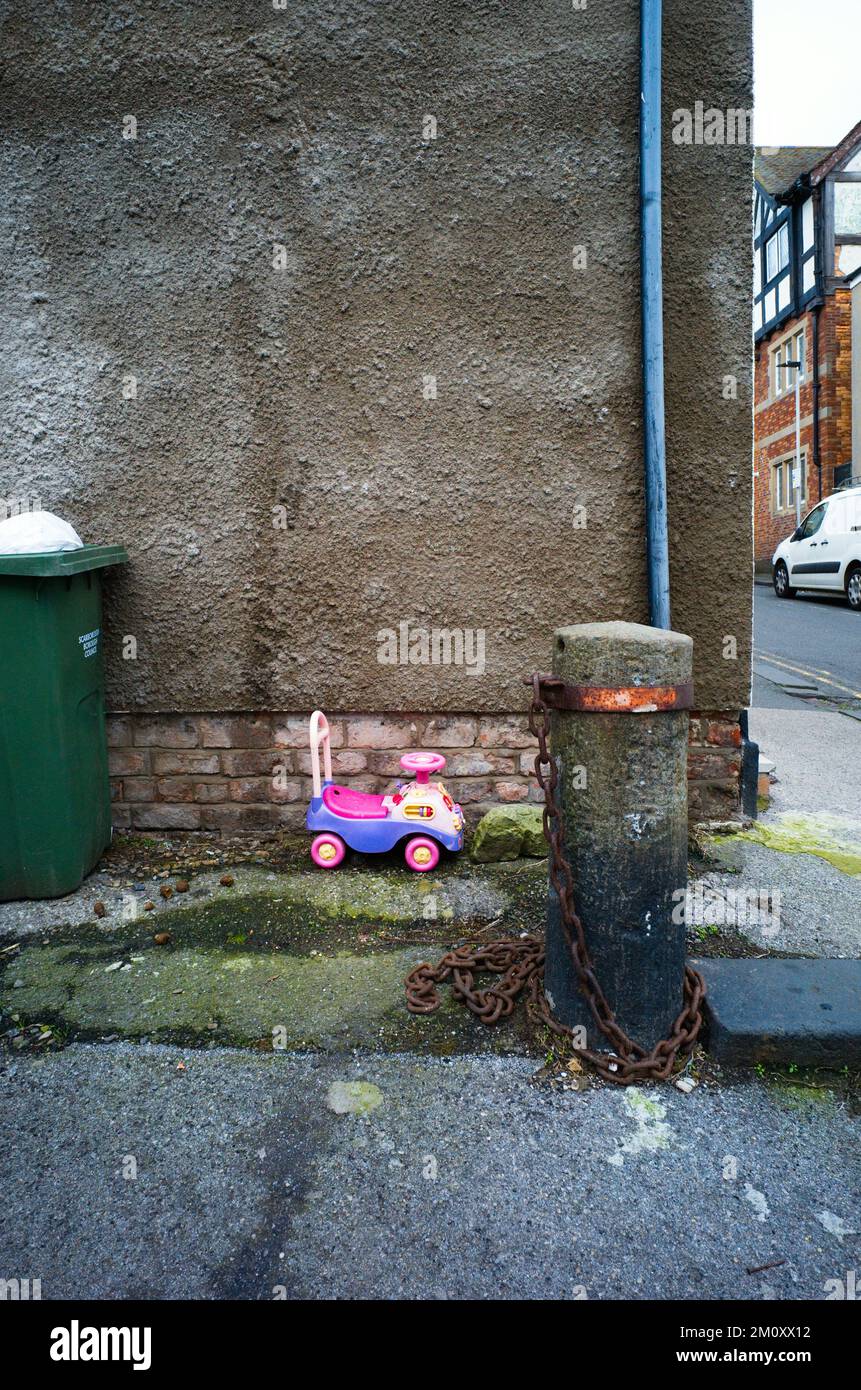 Verlassene Kinder sitzen auf dem Spielzeug neben dem Mülleimer und dem angeketteten Pfosten Stockfoto