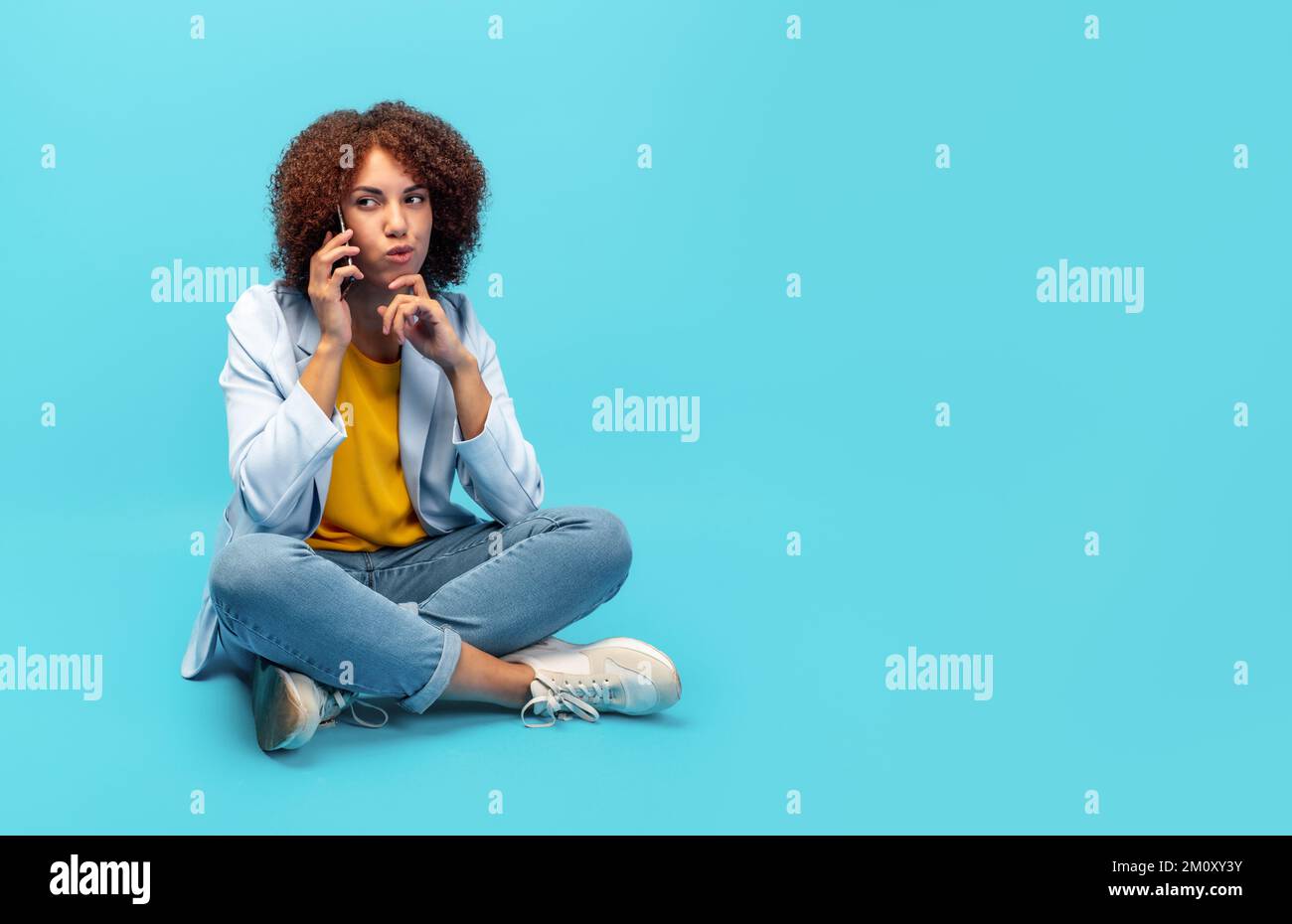 Junge afroamerikanische Frau in Jacke, die mit dem Handy spricht, während sie auf blauem Hintergrund sitzt Stockfoto
