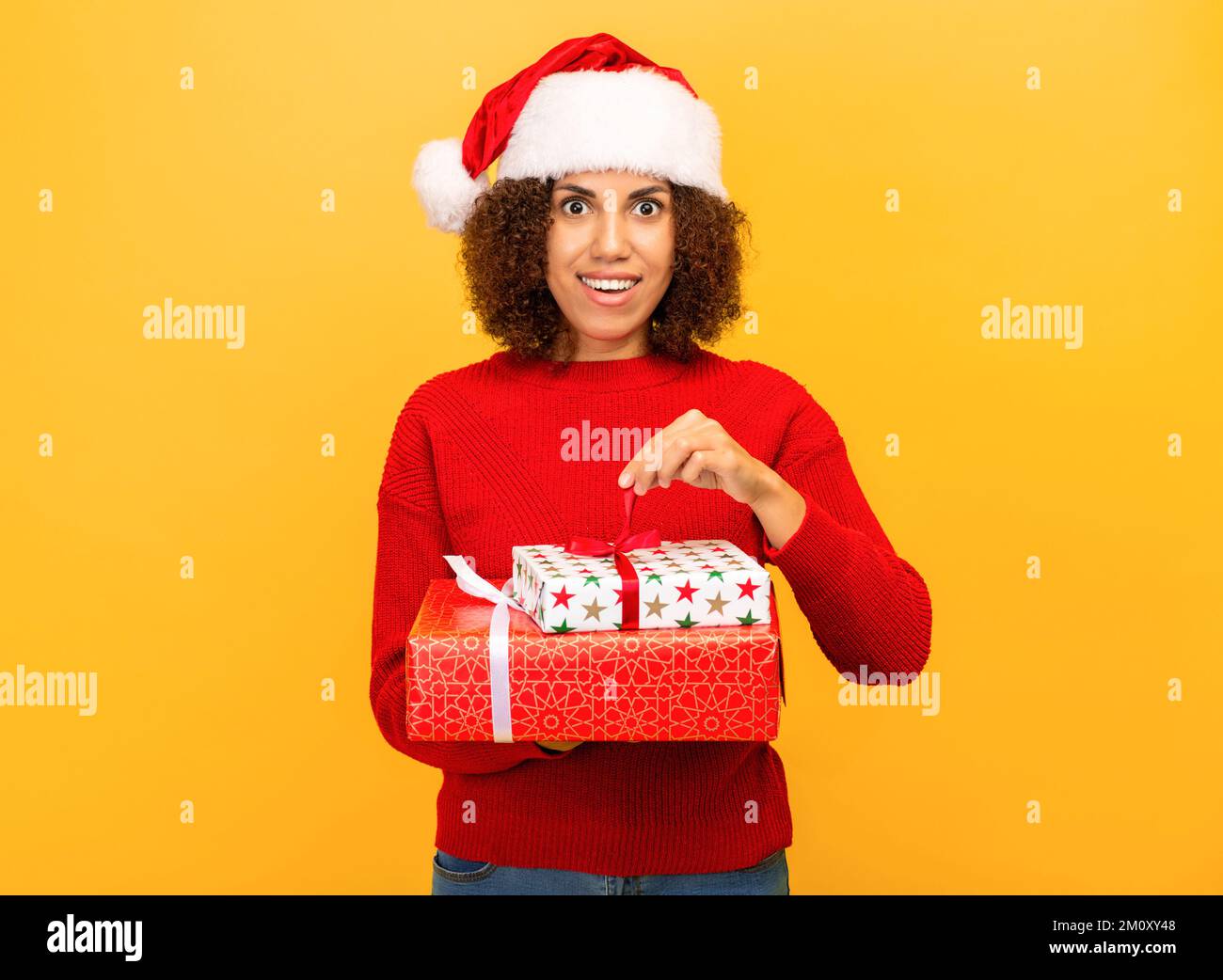 Angenehm aufgeregte glückliche Frau hält ein weihnachtsgeschenk in Händen. Frau mit weihnachtsmann-Hut auf orangefarbenem Hintergrund, lächelnd Stockfoto