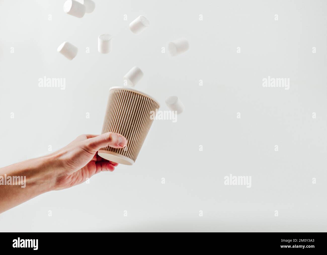 Pappbecher mit heißem Getränk, fliegende Marshmallows in männlicher Hand. Weißer Hintergrund. Kaffee, Latte, Cappuccino, Kakao. Einweg-Pappbecher. Recycling und e Stockfoto