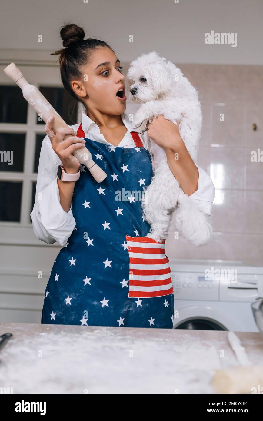 Eine fröhliche Frau mit einer rollenden Nadel, die einen süßen, weißen Malteser in der Küche hält Stockfoto