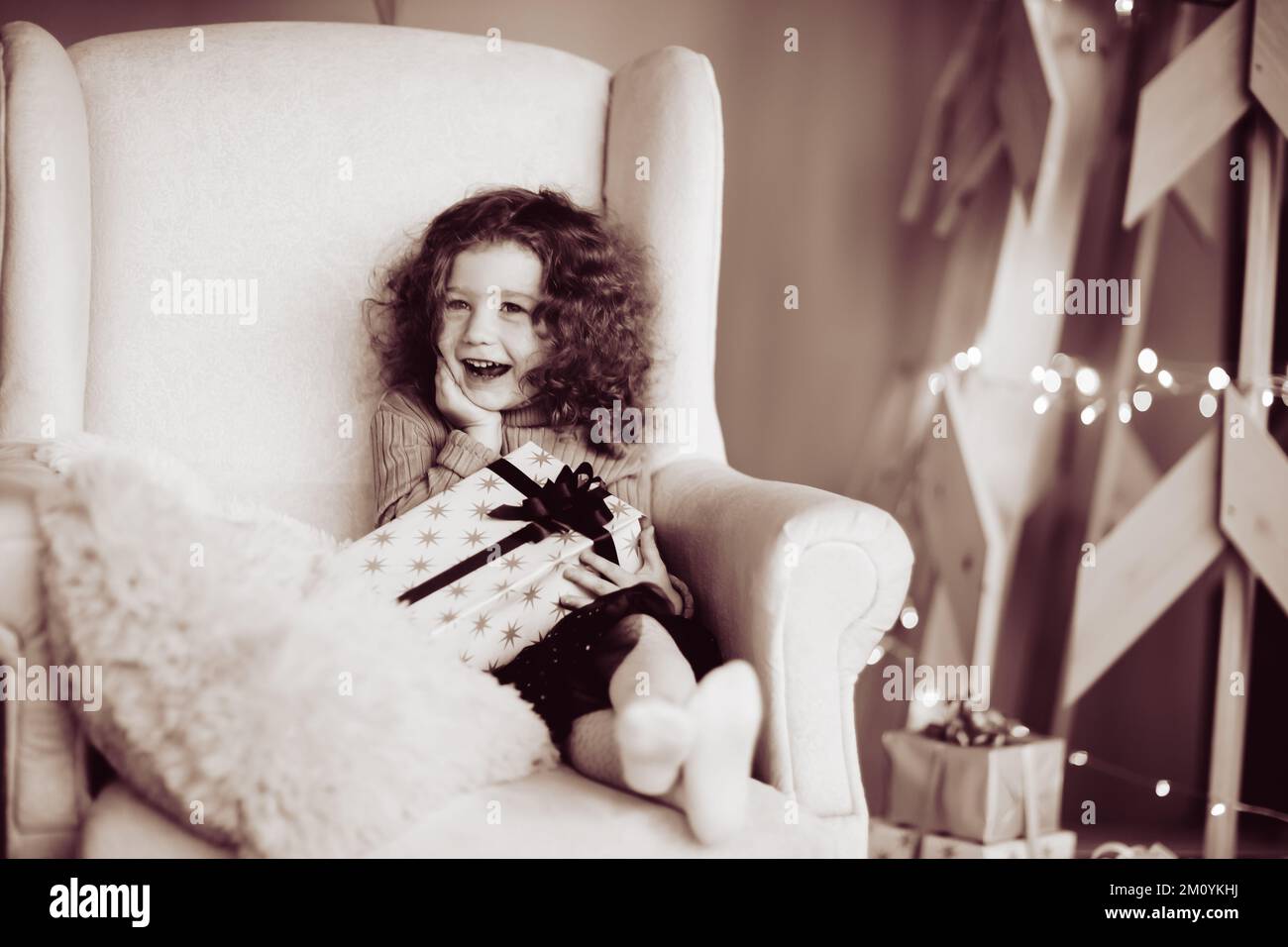 Glückliches, süßes Mädchen mit einem Weihnachtsgeschenk, das in einem gemütlichen Stuhl sitzt. Schwarzweißfoto Stockfoto