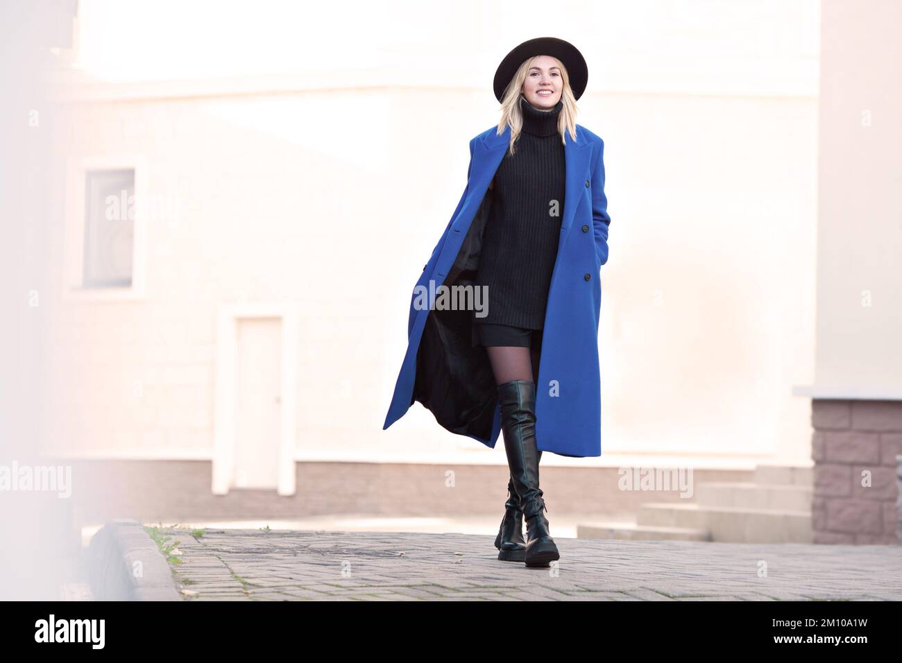 Fröhliches Model mit einem schönen Lächeln, das die Straße in Bewegung hält. Blonde Frau mit Fedora, blauem Herbst- oder Frühlingsmantel, schwarzem Strickpullover, Shorts, Hig Stockfoto