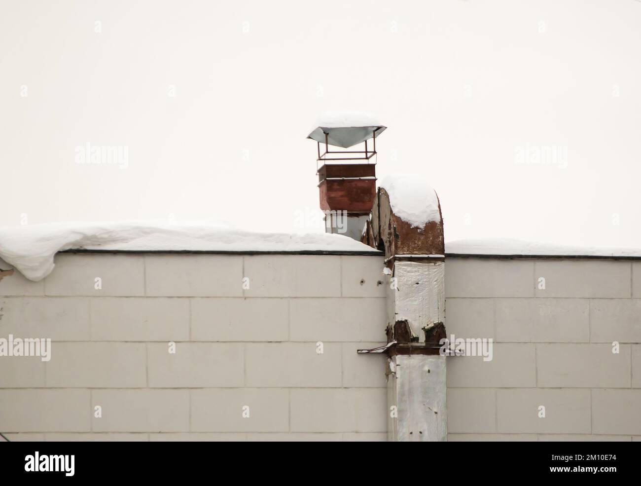 Rostiges Vierkantrohr aus Metall für Abluft und Belüftung an der Hauswand. Das Dach des Gebäudes in weißem Schnee. Bewölkt, kalter Wintertag, weiches Licht. Stockfoto