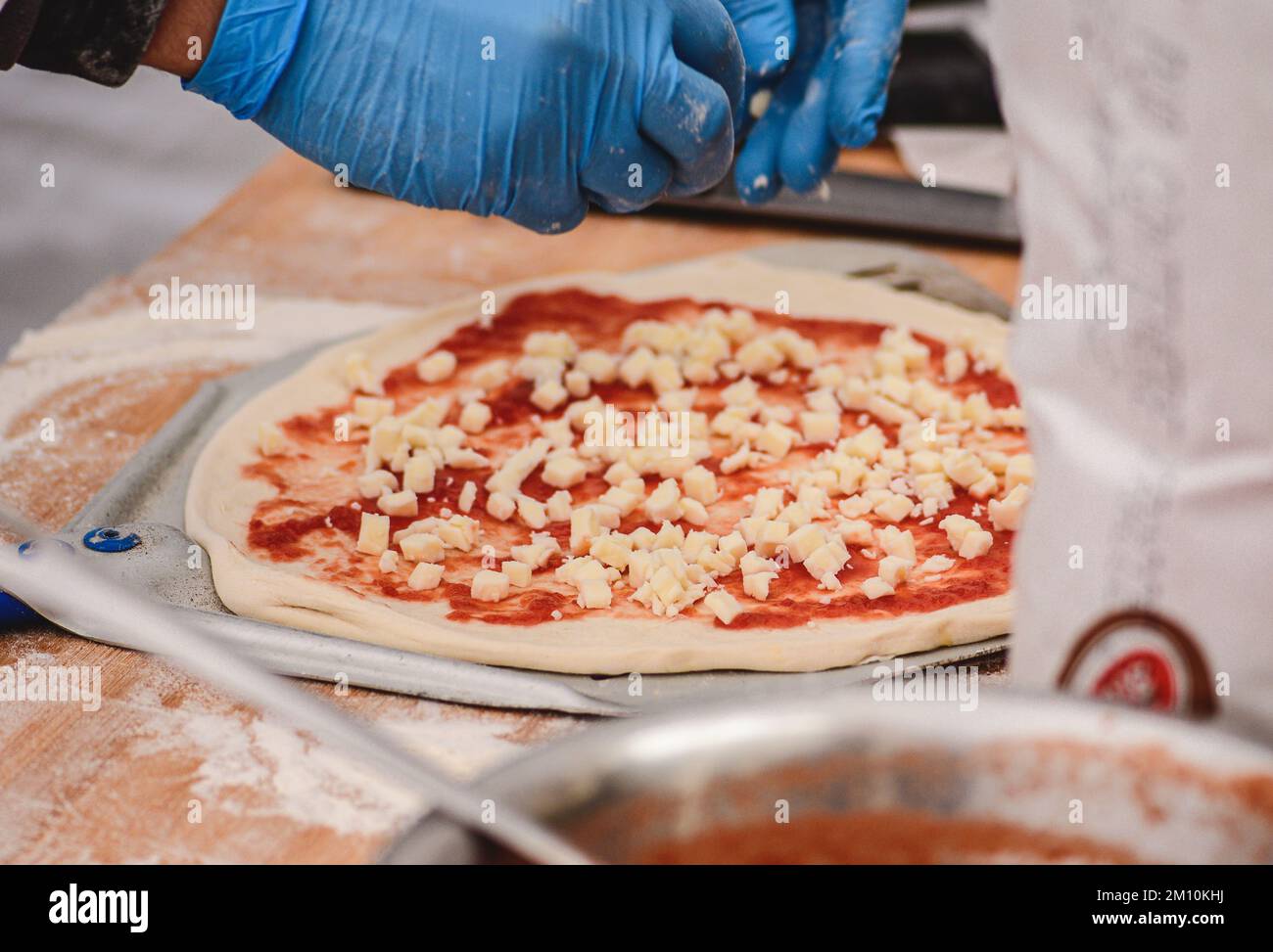 Hände mit Kunststoffhandschuhen Fügen Sie frischen Mozzarella auf eine Pizza mit Tomatensoße auf einem Holzbrett mit Zutaten hinzu, Nahaufnahme Stockfoto