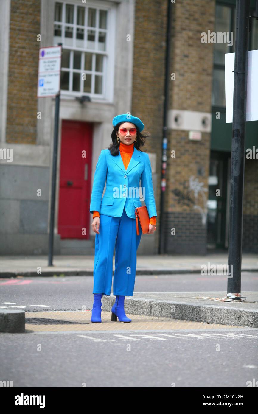 Schicke, stilvolle Frau mit blauer Baskenmütze, blauen Stiefeln, blauen Hosen, blauer Jacke, orangefarbenem Rollkragenpullover, Eine Sonnenbrille und eine orangefarbene Unterarmtasche in einer Londoner Straße Stockfoto