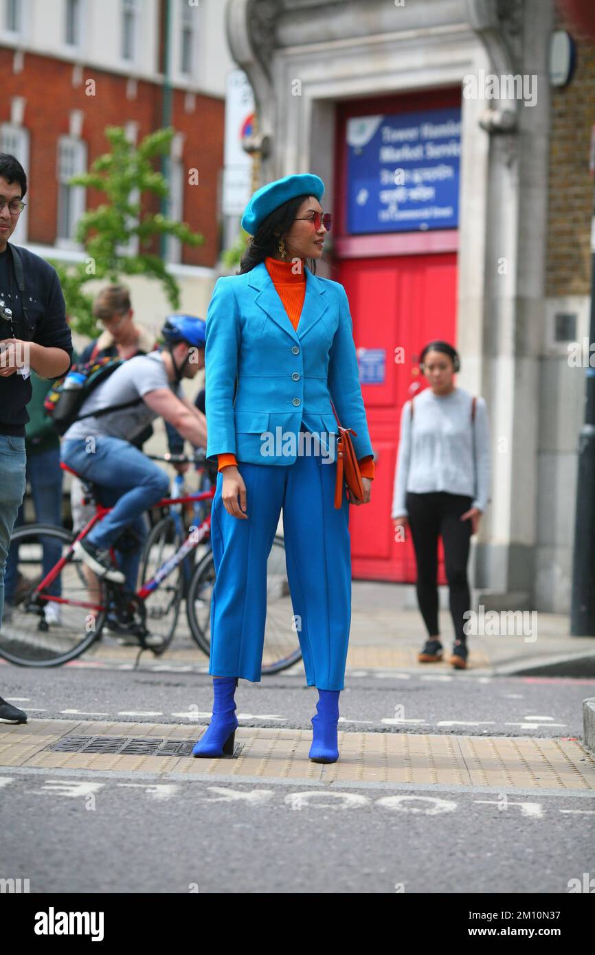 Schicke, stilvolle Frau mit blauer Baskenmütze, blauen Stiefeln, blauen Hosen, blauer Jacke, orangefarbenem Rollkragenpullover, Eine Sonnenbrille und eine orangefarbene Unterarmtasche in einer Londoner Straße Stockfoto