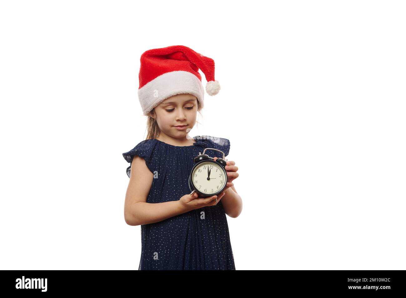 Hübsches kleines Mädchen mit Weihnachtsmannmütze und Abendkleid, mit schwarzem Retro-Wecker, der Mitternacht anzeigt. Weihnachten Stockfoto