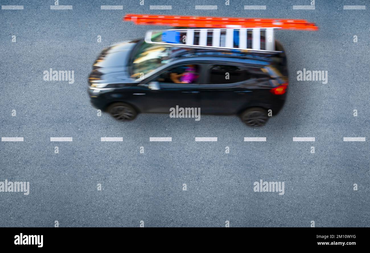 Draufsicht eines Fahrers, der in einem kompakten schwarzen Auto mit einer orangefarbenen Trittleiter arbeitet, die am Dachgepäckträger befestigt ist. Stockfoto
