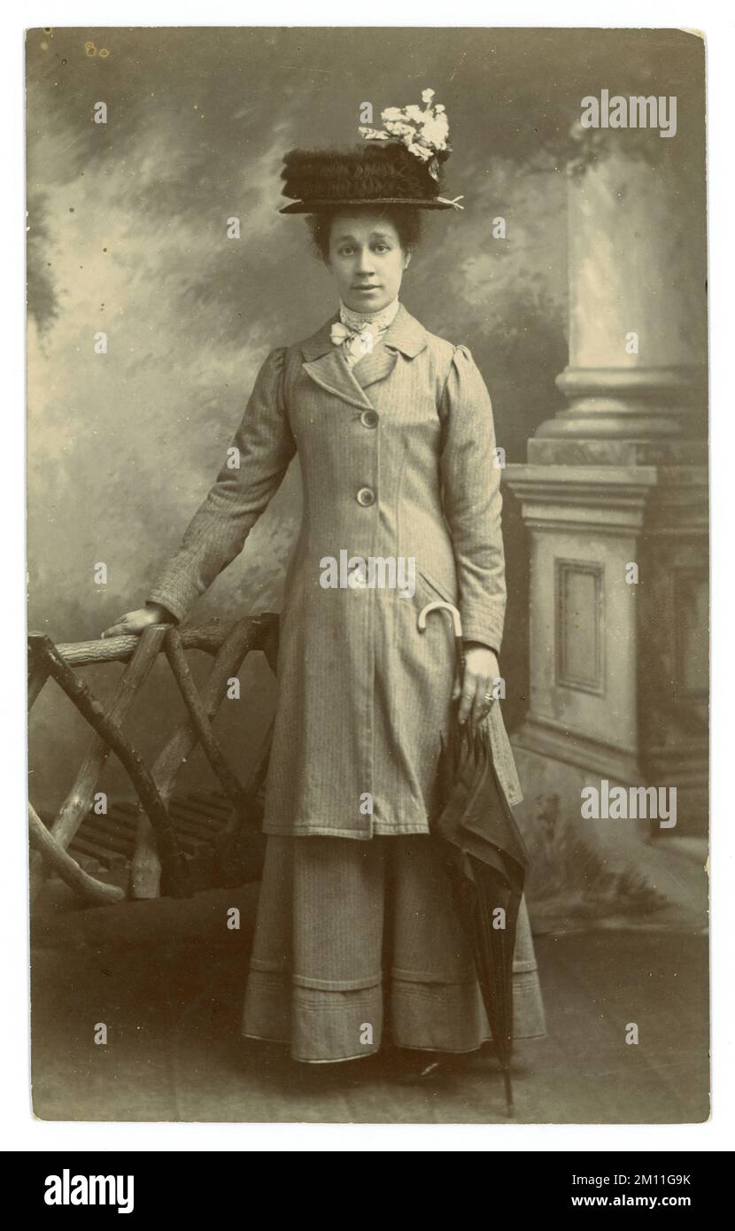 Original Edwardians Atelierpostkarte mit attraktiver Frau in langer Jacke und Rock, großem Hut mit Rüschenspitze und Blumenzweig, mit einem Regenschirm mit Stock Griff. Edwardianerin. Edwardianerin. Juli 1910, Großbritannien Stockfoto