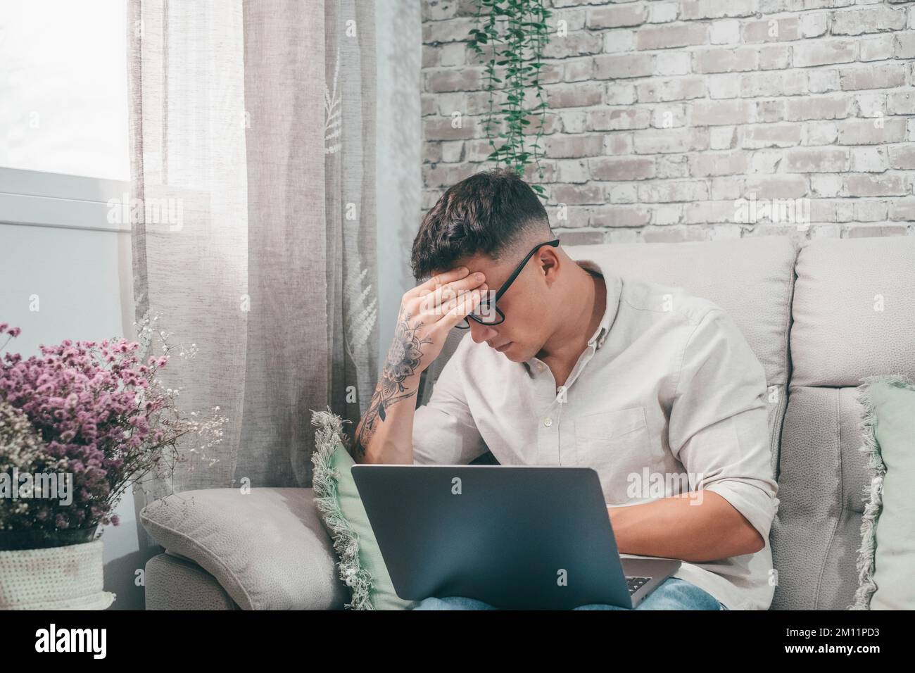 Ein junger kranker Mann, der auf dem Sofa sitzt und sich schlecht fühlt, mit Schmerzen und Kopfschmerzen. Ein Teenager mit einer Tätowierung am Arm, der sein Gesicht berührt, ist krank. Intellektueller Teenager mit Brille. Stockfoto