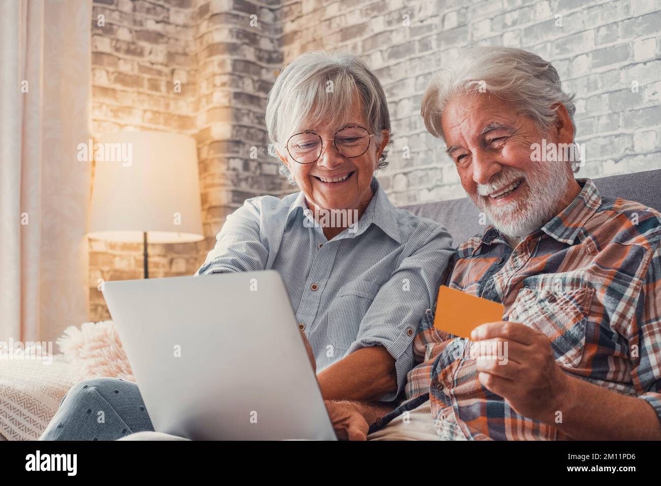 Porträt von zwei süßen, glücklichen und aufgeregten Senioren, die zu Hause mit einem Laptop auf dem Sofa online einkaufen und Geld ausgeben. Gemeinsame Nutzung von Kreditkarten beim Kauf im Internet. Stockfoto
