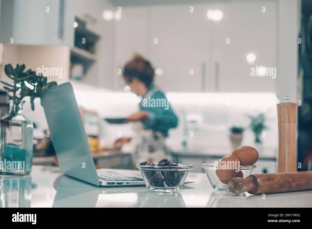 Im Vordergrund stehen Laptop und Lebensmittelzutaten in der Küche zu Hause. Eine Frau im Hintergrund kochte unscharf und genoss die Mittagsvorbereitung allein. Realer Lifestyle und moderne Online-Inhalte Stockfoto