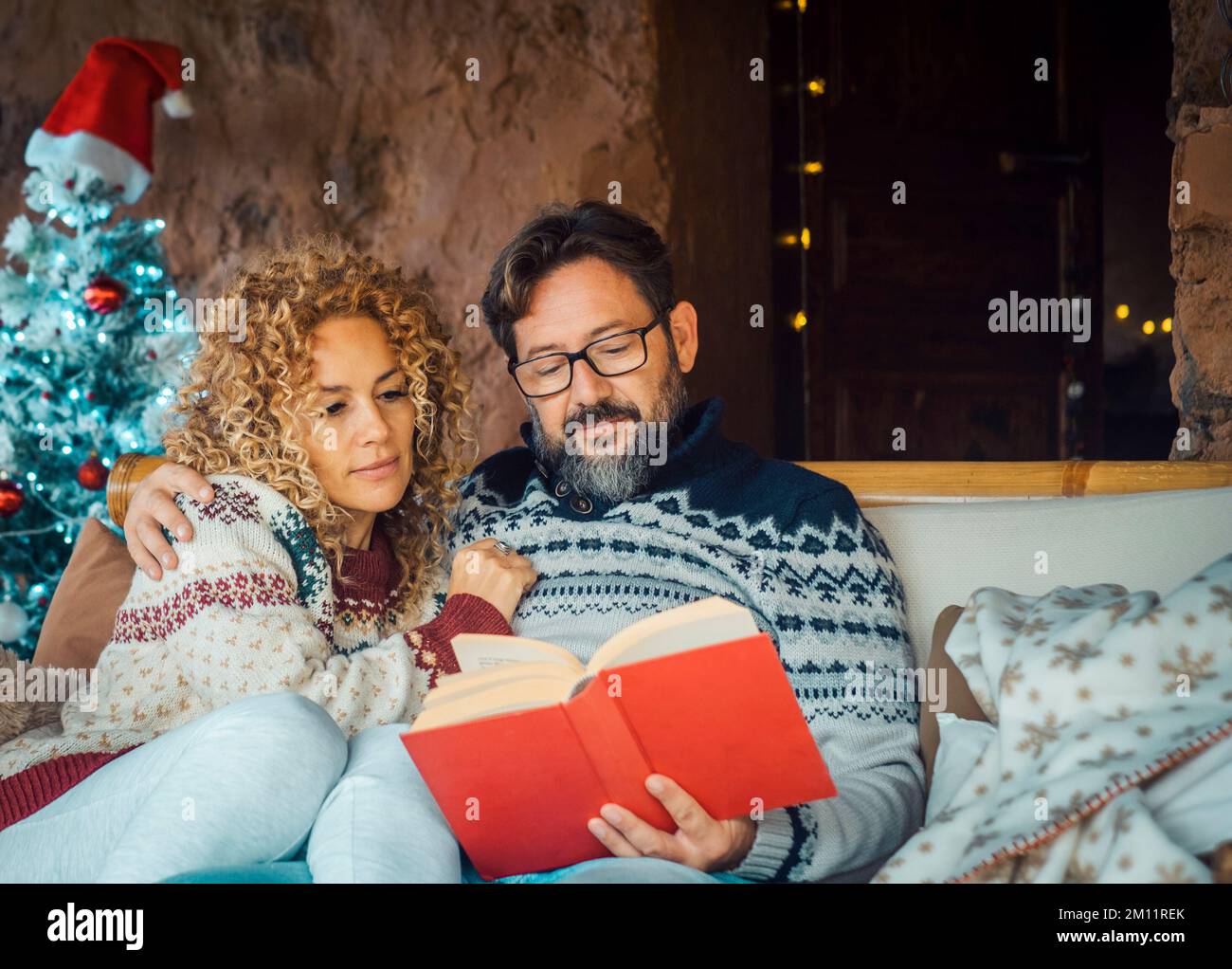 Mann und Frau zu Hause genießen die Feiertage und die weihnachtszeit bei entspannter Freizeitbeschäftigung im Innenbereich. Menschen in einer Beziehung. Ein erwachsener Mann liest ein Buch. Frau umarmt Ehemann und lächelt Stockfoto