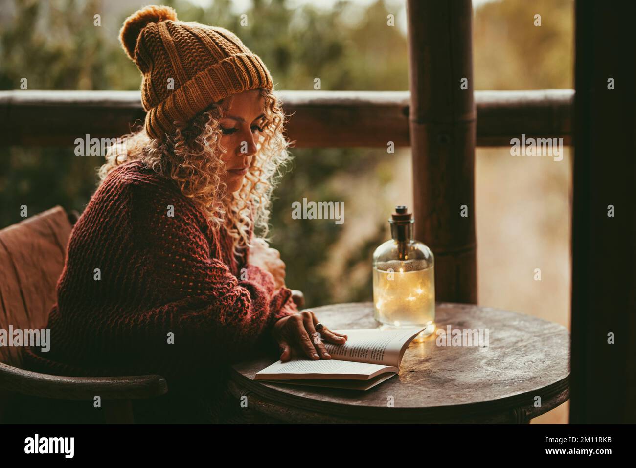 Eine Frau, die draußen ein Buch liest, sitzt auf einem kleinen Holztisch und trägt Pullover und Wollmütze für kalte Temperaturen. Weibliche Erwachsene und Erziehung in Freizeitbeschäftigung zu Hause. Allein die hübsche Dame Stockfoto