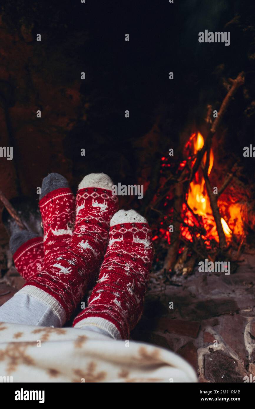 Ein paar Füße wärmend neben dem Kamin zu Hause oder im Chalet-Zimmer. Urlaub in der weihnachtszeit. Romantische Menschen genießen Beziehungen und Zärtlichkeit, die drinnen wie ein Feuer aussehen. Weihnachtssocken in Rot und Weiß Stockfoto