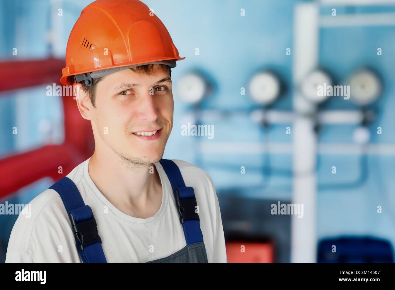 Männliches Porträt eines Bauhelmarbeiters im Industriebau mit Drucksensoren und Druckmessern im Hintergrund. Junger Mann in Arbeitskleidung lächelt und schaut in die Kamera. Stockfoto