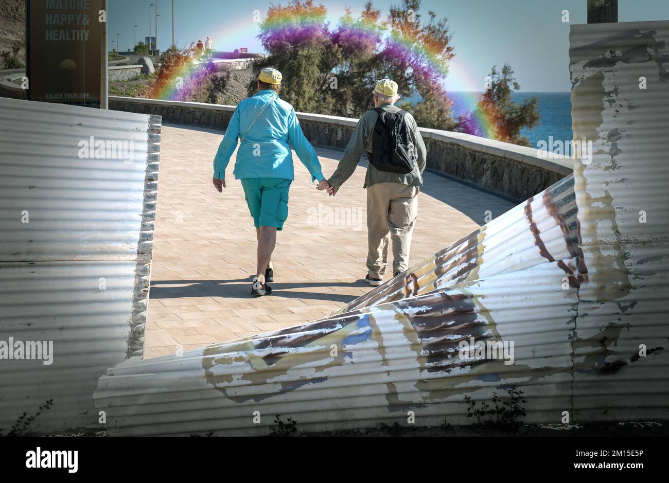 Ein pensioniertes Paar, das Hand in Hand auf der Promenade in Richtung eines Regenbogens (Verbundwerkstoff) spaziert. Stockfoto