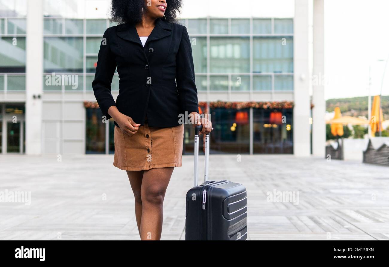 Pflückende afroamerikanische Geschäftsfrau mit passender schwarzer Jacke und lächelndem Lächeln, während sie tagsüber auf der Straße spaziert Stockfoto