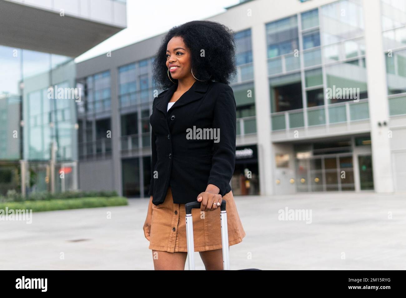 Glückliche afroamerikanische Geschäftsfrau mit Koffer, die die schwarze Jacke anpasst und lächelt, während sie tagsüber auf der Straße der Stadt spaziert Stockfoto