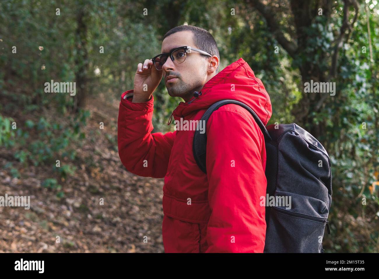 Seitenansicht eines jungen Mannes in roter Jacke und Rucksack mit stilvollen Gläsern und Bewunderung des Waldes am Wochenende auf dem Land Stockfoto