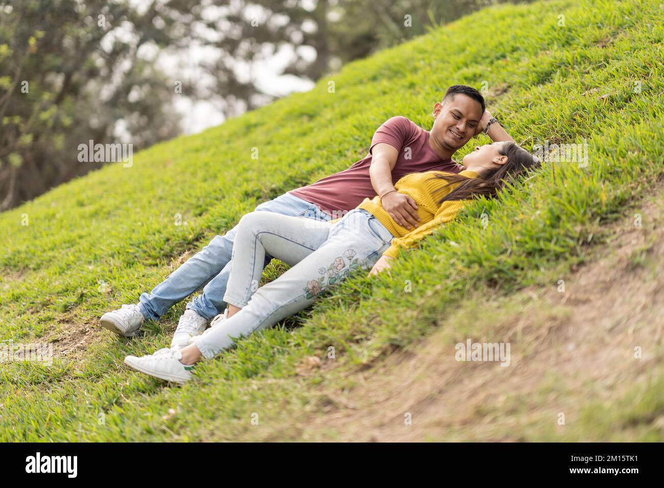 Fröhlicher junger Mann und Frau in lässiger Kleidung, lächelnd und schauend sich an, während sie sich während eines romantischen Dates auf dem Gras im Park entspannen Stockfoto