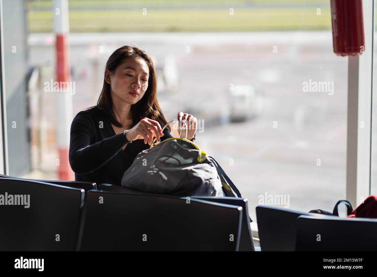 Junge asiatische Frau mit dunklem Haar nimmt Draht aus der Tasche, während sie tagsüber auf einem Stuhl am Fenster im Terminal des modernen Flughafens sitzt Stockfoto