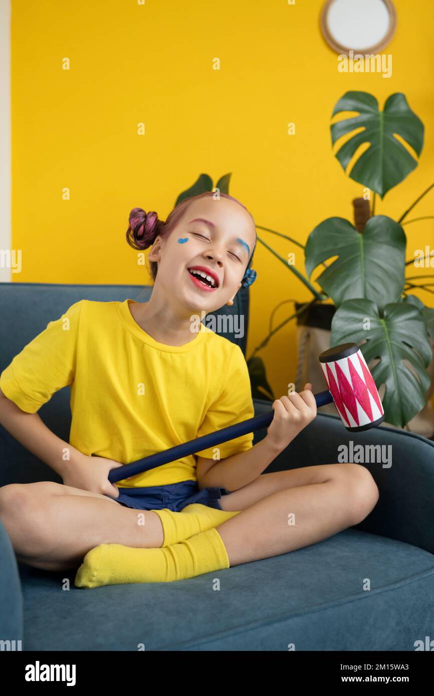 Der ganze Körper eines positiven kleinen Mädchens mit pinkfarbenen und blauen Brötchen und Make-up sitzt im Studio auf einem Sessel mit gekreuzten Beinen und Cospying Comic-Figur Stockfoto