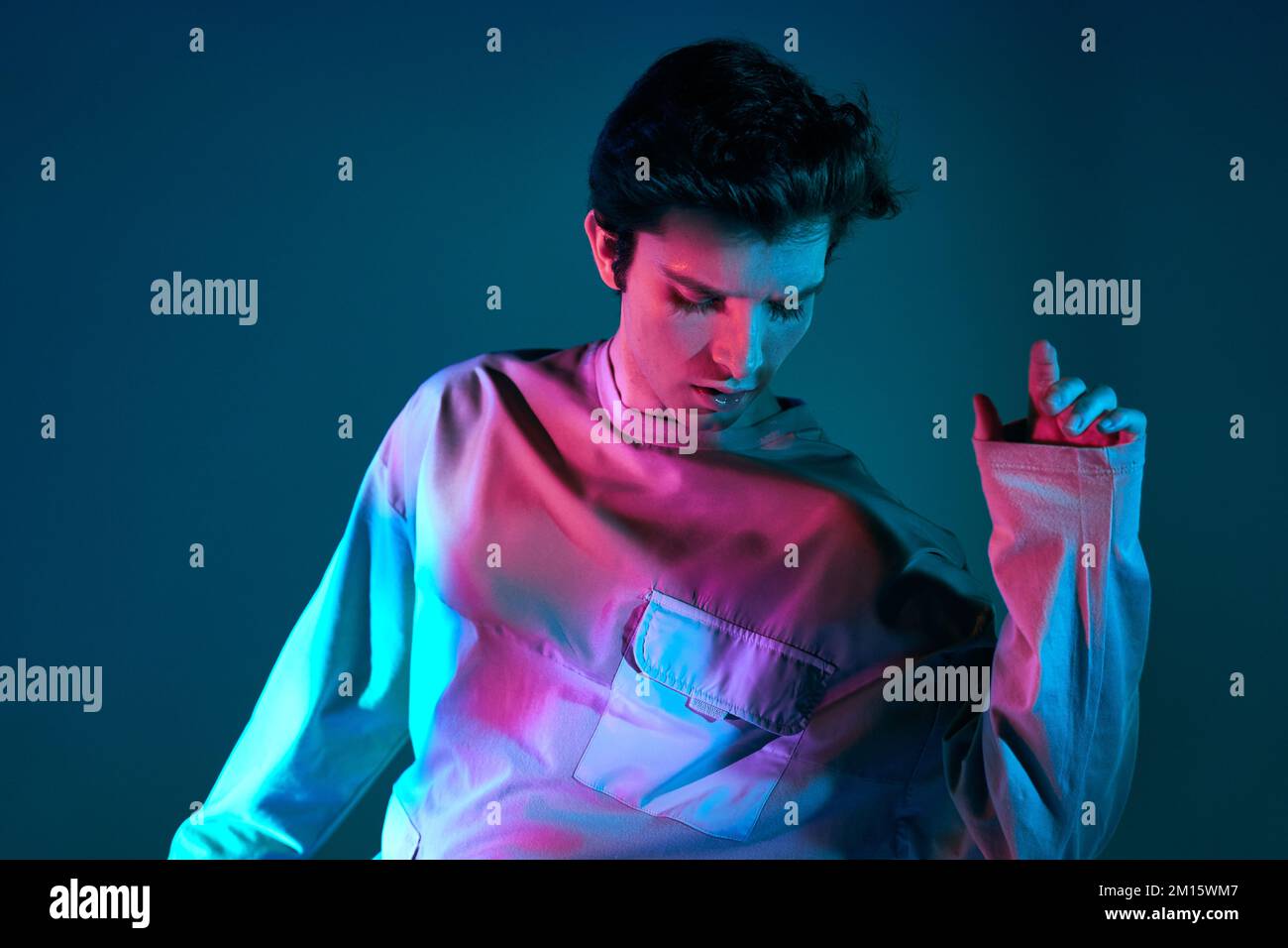 Junger Mann, dunkelhaariges Modell in Sweatshirt mit vielen Taschen, steht im Studio mit geschlossenen Augen in leuchtenden, mehrfarbigen Neonlichtern Stockfoto